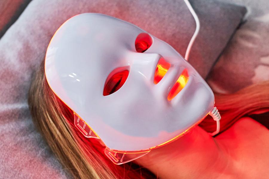 a person in a robe sitting on a couch reading a magazine with an LED face mask on