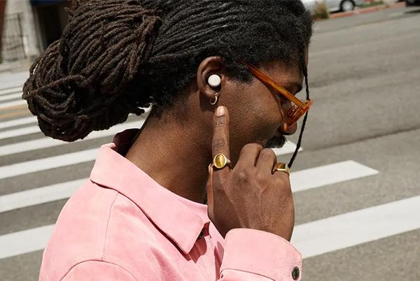 a man with dreadlocks wearing a pink shirt and pointing towards google pixel buds pro 2 earbud in his ear