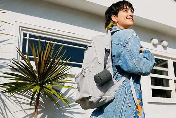 a woman in a denim jacket and backpack walking down the street with sony srs-xb100 portable speaker