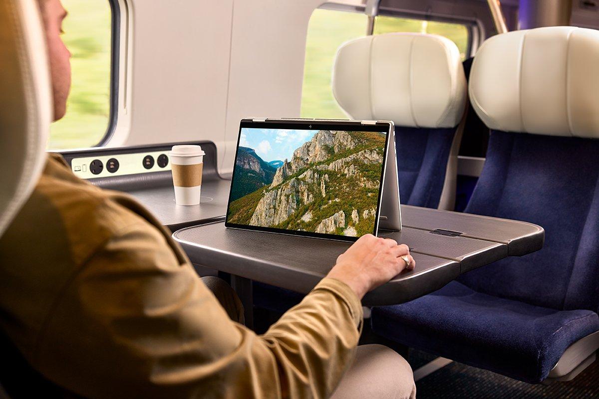 A man sits on a train with an HP Envy 16 on the table in front of him
