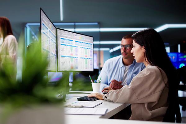 a man and woman sitting at a desk looking at a dual monitor desktop computer