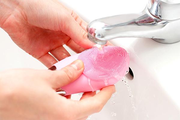 a person washing their silicone face cleansing brush under a tap in a sink