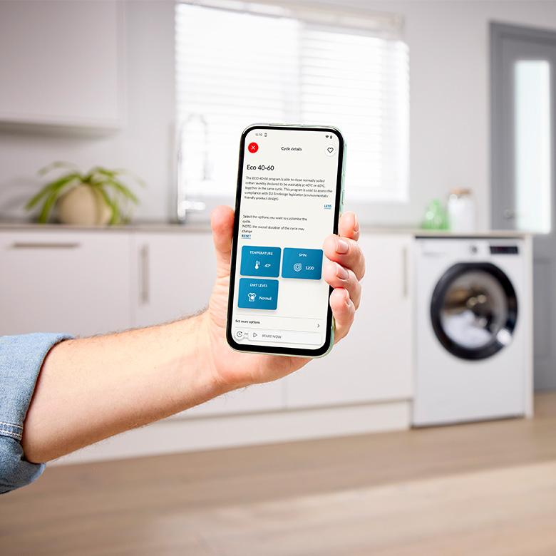 a person holding up a smart phone in a kitchen with hoover h-wash 350 wi-fi washing machine in background
