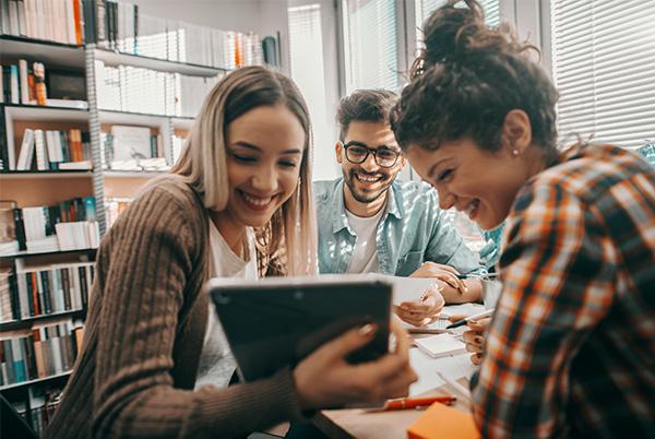 three young people sitting at a table in a library looking at a tablet