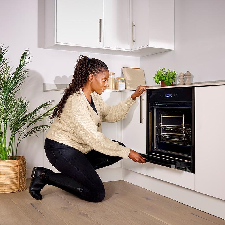 a woman kneeling down in front of a neff n70 slide and hide electric pyrolytic oven opening it