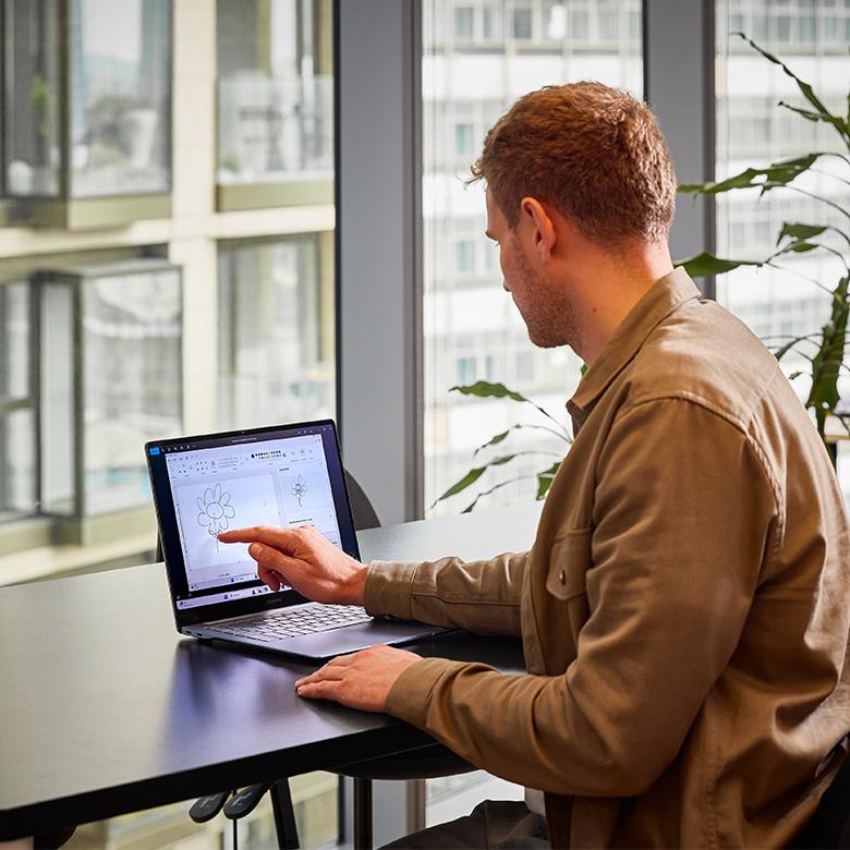 a man sitting at a desk with a laptop in front of him using his finger to draw a flower on the touchscreen