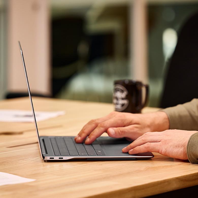 a person typing on a thin laptop computer at a table with a cup of coffee