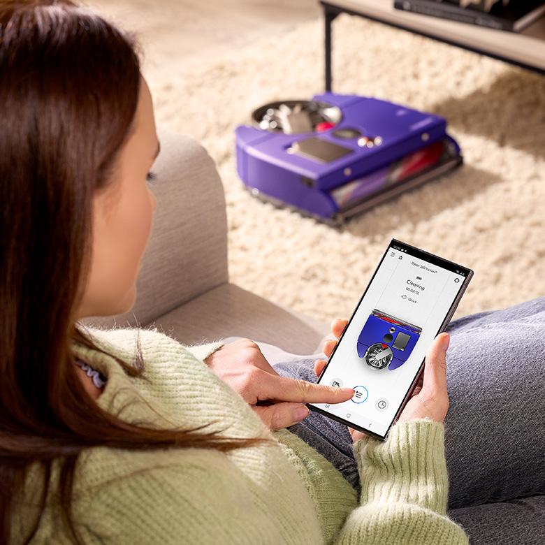 a woman sitting on a couch holding her phone using mydyson app to control 360 vis nav robot vacuum cleaner on floor