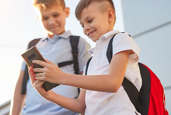 Two boys with backpacks standing next to each other looking at a mobile phone.