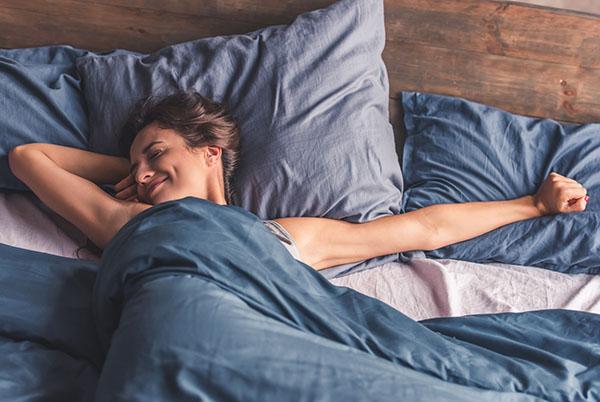 A relaxed woman laying in bed under a weighted blanket, with arms outstretched and smiling