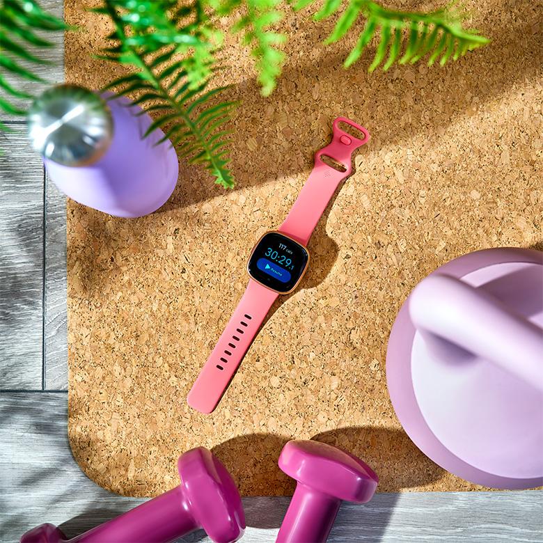 a pink fitbit versa 4 smart watch on a table next to weights and a plant