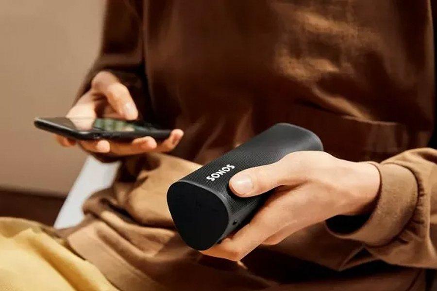 A person holding a mobile phone in one hand and a Sonos speaker in the other