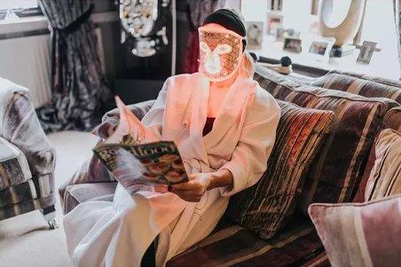 a person in a robe sitting on a couch reading a magazine with an LED face mask on