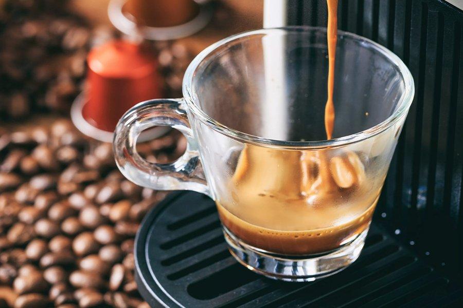 A glass cup on a black tray with coffee pouring into it