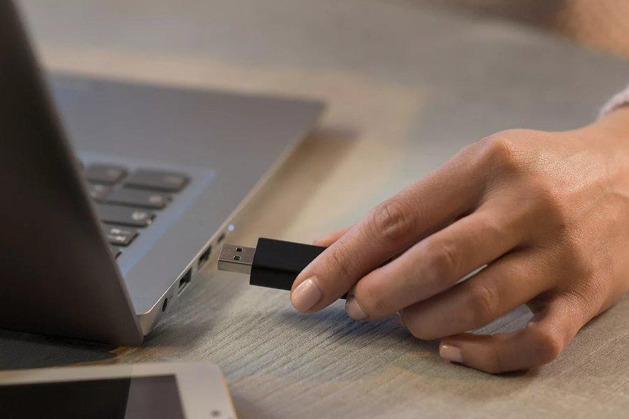 A hand holding a usb flash drive next to a laptop