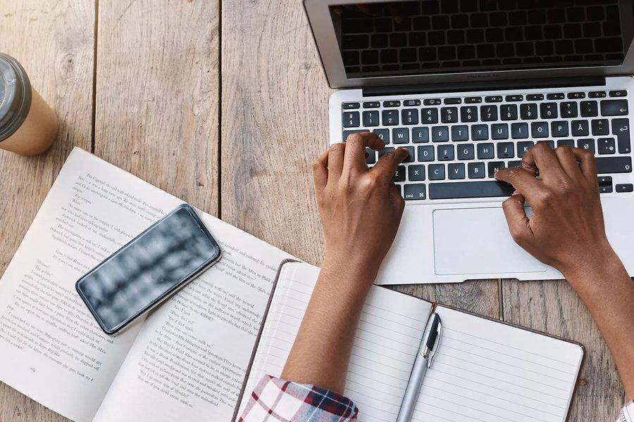 A pair of hands on a laptop with a book, notebook and mobile phone next to it