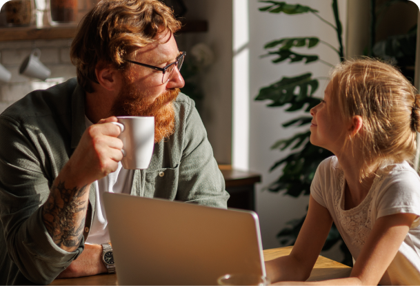 A father and daughter sit at a table with a laptop on it, the father is drinking from a mug 