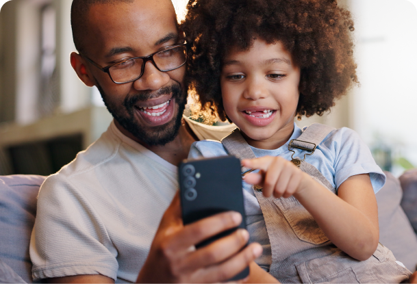 A son on sitting on his father whilst they both look at a mobile phone