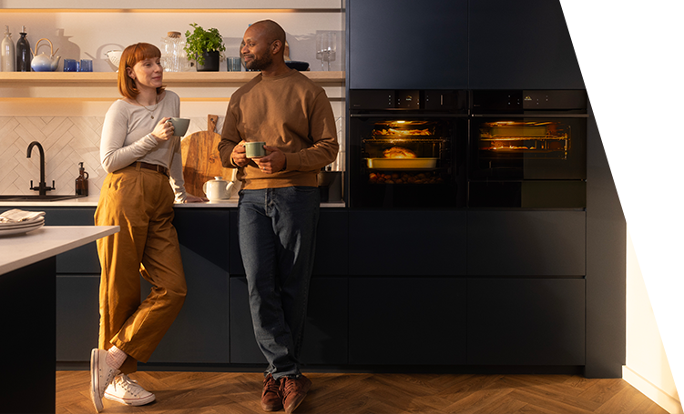 A man and a woman standing in a kitchen next to two built-in ovens
