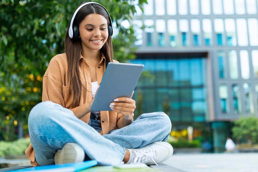 A woman sitting outside using a tablet whilst wearing headphones
