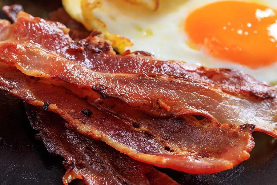 A close up of some crispy bacon with a fried egg in the background
