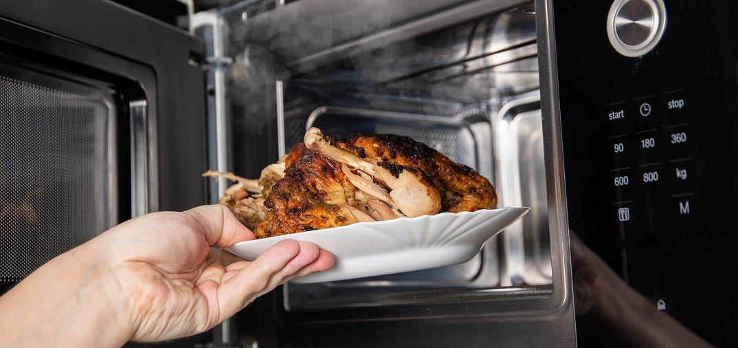 a person holding a cooked chicken in a white dish in front of a microwave