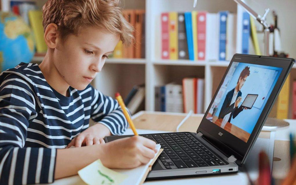 A boy writing on a notepad whilst using a laptop