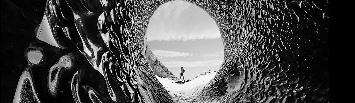 Black and white image of a person trekking in the distance from the perspective of a cave showing dazzling darks and luminous lights