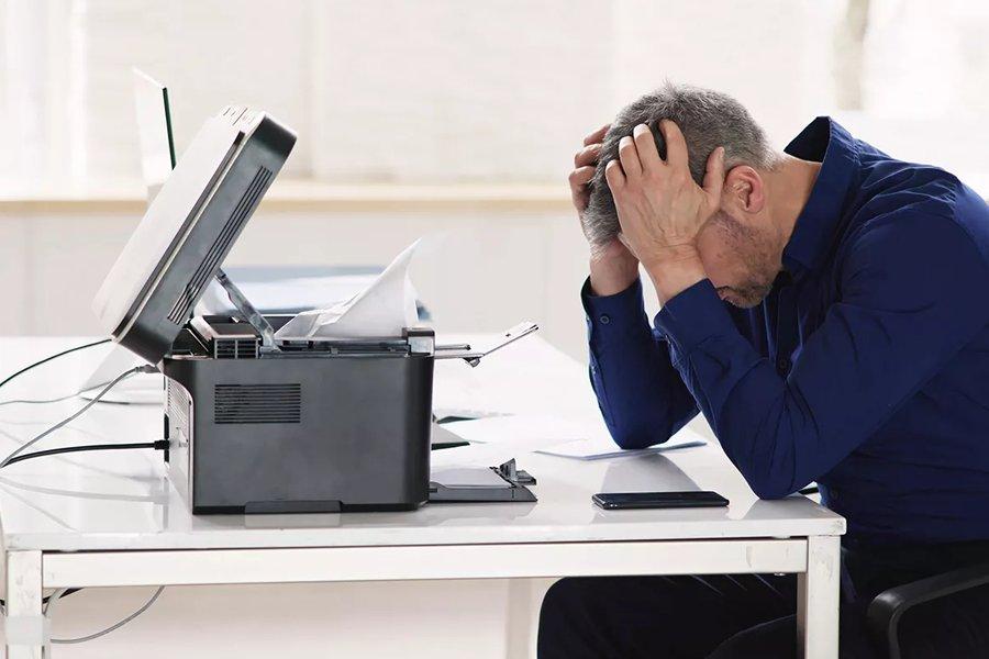 A man sitting at a desk with his head in his hands with a broken printer in front of him