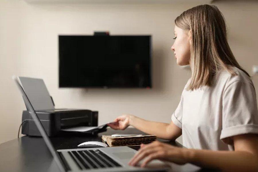 A woman sitting at a desk reaching out to a printer with a laptop in front of her
