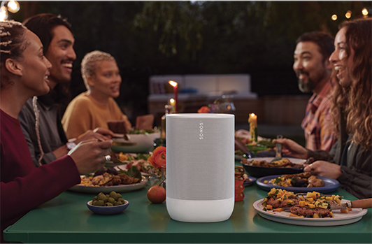 A white speaker is on a table, people are sitting around it talking