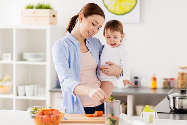 a woman holding a baby in a kitchen with vegetables