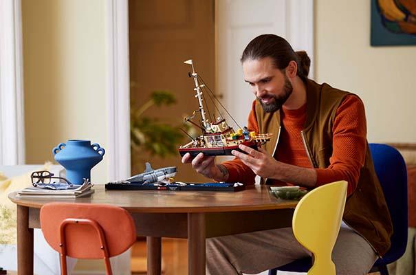 a man sitting at a table playing with a lego ship