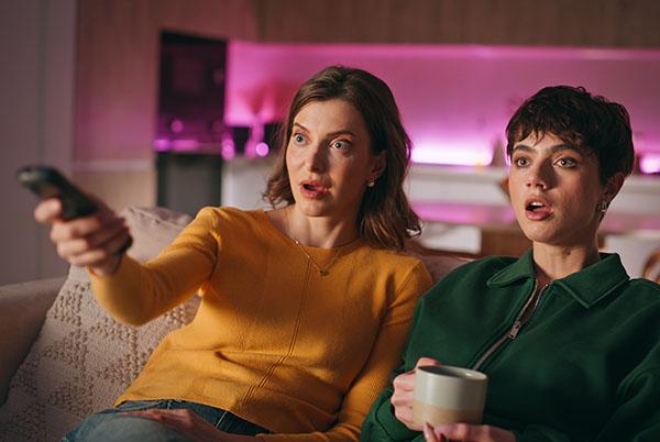two women sitting on a couch watching tv with a cup of coffee