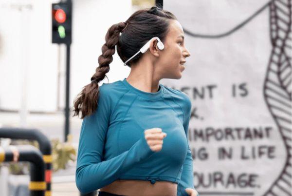 a woman in a blue shirt running with a white Shokz OpenMove bone conducting headphones on her ear