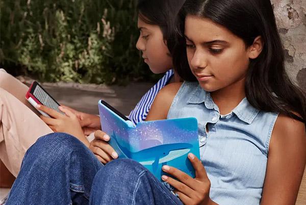 two girls sitting on a bench reading books on their tablets