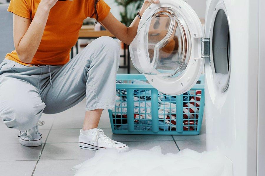 A person crouched next to an open washing machine with bubbles all over the floor