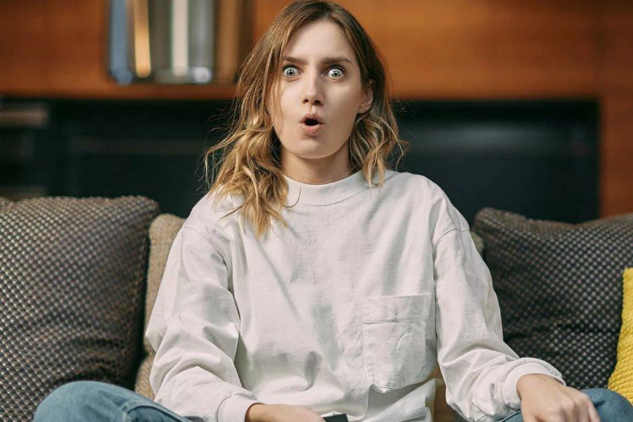 A surprised woman sitting on a coach while watch TV